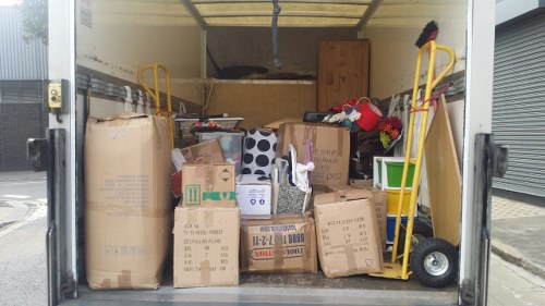 Inside a commercial van used for rubbish removal, there is a dense collection of cardboard boxes, some flattened and others partially intact, of varying sizes and brown in colour, with printed labels and logos. Among the boxes, there are plastic containers and storage bins, including a white one with a green handle, and a black-and-white patterned bag. A large beige package or bag is positioned on the left, partially obscuring the view. Two yellow hand trucks or dollies are situated on either side of the van, leaning against the interior walls, with the one on the right supporting a small black object at the top. The van’s interior floor appears to be covered with miscellaneous waste materials, and the back wall is light-colored with a wooden panel at the far end. Outside, a portion of a paved driveway or pavement is visible to the right, indicating a commercial or industrial setting. The scene suggests a collection of waste items awaiting disposal or collection, representative of a professional rubbish removal service that could operate in North Watford or nearby areas.