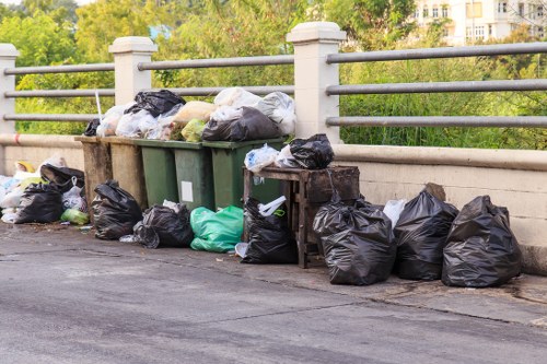 A collection of black and green rubbish bags, along with some white and grey trash sacks, piled on the pavement next to a green waste bin and an orange-brown waste container, situated on a paved area adjacent to a metal railing. The bags are stacked close to the edge of a roadway, with additional bags placed on the ground in front of the containers. In the background, there is a view of trees and multi-storey residential buildings, indicating an urban or suburban environment. The scene suggests a recent rubbish collection or disposal activity, typical of waste removal services in North Watford. The lighting appears natural, with daylight illuminating the scene, emphasizing the textures of the bags and the surrounding environment, suitable for a website offering commercial waste removal in the area.