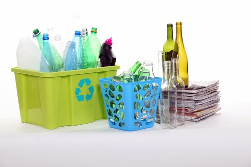 A collection of recyclable waste items arranged on a plain white surface, including two plastic crates—one lime green with a recycling symbol and one blue—overflowing with empty plastic bottles of various colours such as clear, green, and blue, as well as glass bottles in shades of green and amber. Several glass bottles, some tall and slender, others shorter and wider, are standing upright next to a neat stack of newspapers or magazines. The background is plain and uncluttered, emphasizing the different materials and their textures, with the bottles displaying smooth, shiny surfaces and the paper having a matte finish. This scene reflects a typical setup for a waste collection or recycling process, highlighting the importance of separating different waste streams for effective rubbish removal, similar to services offered by Commercial Waste North Watford in the local area. The overall composition presents an organized and clean display of recyclable waste destined for proper disposal or recycling in the North Watford region.