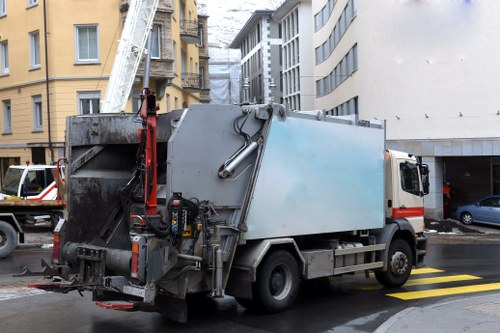A medium-sized commercial waste collection truck parked on a city street, with a large rectangular metal waste storage compartment painted in light blue on its chassis. The truck's rear end features an opening mechanism for waste disposal, with hydraulic arms and support structures visible. In the background, there are surrounding multi-storey buildings with brick and concrete facades, some with windows and balconies, indicating an urban environment. The street surface includes yellow pedestrian crossing stripes, and other parked vehicles such as cars are visible nearby. The sky is overcast, suggesting typical weather conditions for North Watford. The scene depicts routine rubbish collection activities in a densely built-up area, emphasizing the company's capacity for commercial waste removal, with the vehicle positioned close to the kerb to facilitate waste loading or disposal.