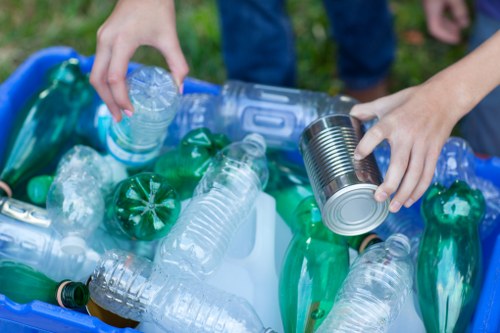 A close-up view of a person's hands placing a clear plastic water bottle into a blue recycling bin filled with various plastic bottles, including green and transparent containers, on a grassy outdoor area. The person is wearing dark jeans, and the background shows an out-of-focus natural setting. The scene illustrates waste separation and recycling efforts, which are part of rubbish removal services provided by Commercial Waste North Watford. The image emphasizes the importance of correctly discarding plastic waste to maintain environmental standards in North Watford and surrounding areas.
